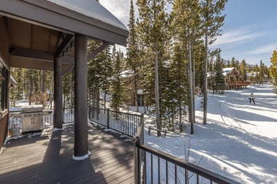 Covered deck with BBQ grill overlooking snowy forest where skiers glide past through the trees.