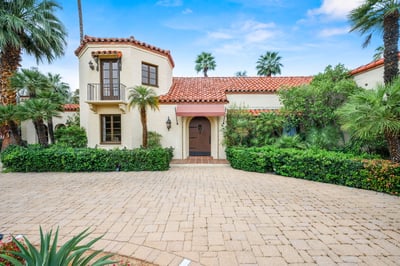 A charming Spanish-style villa with terracotta roof and lush palm gardens framed by a sunny blue sky